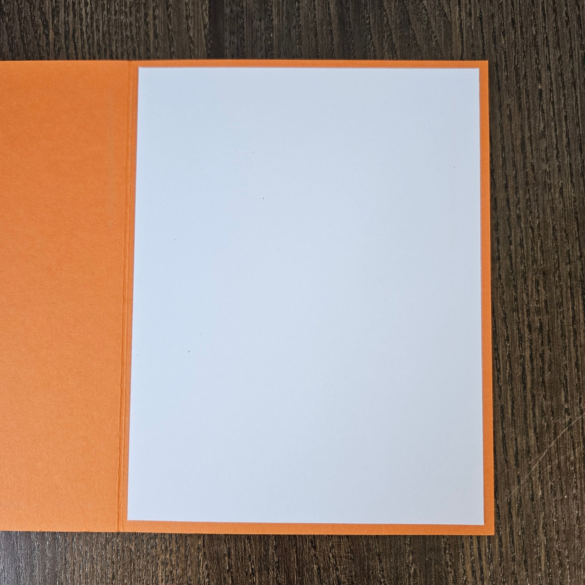 An open orange greeting card or notebook with a blank white page, photographed from above on a dark wood-grain surface.