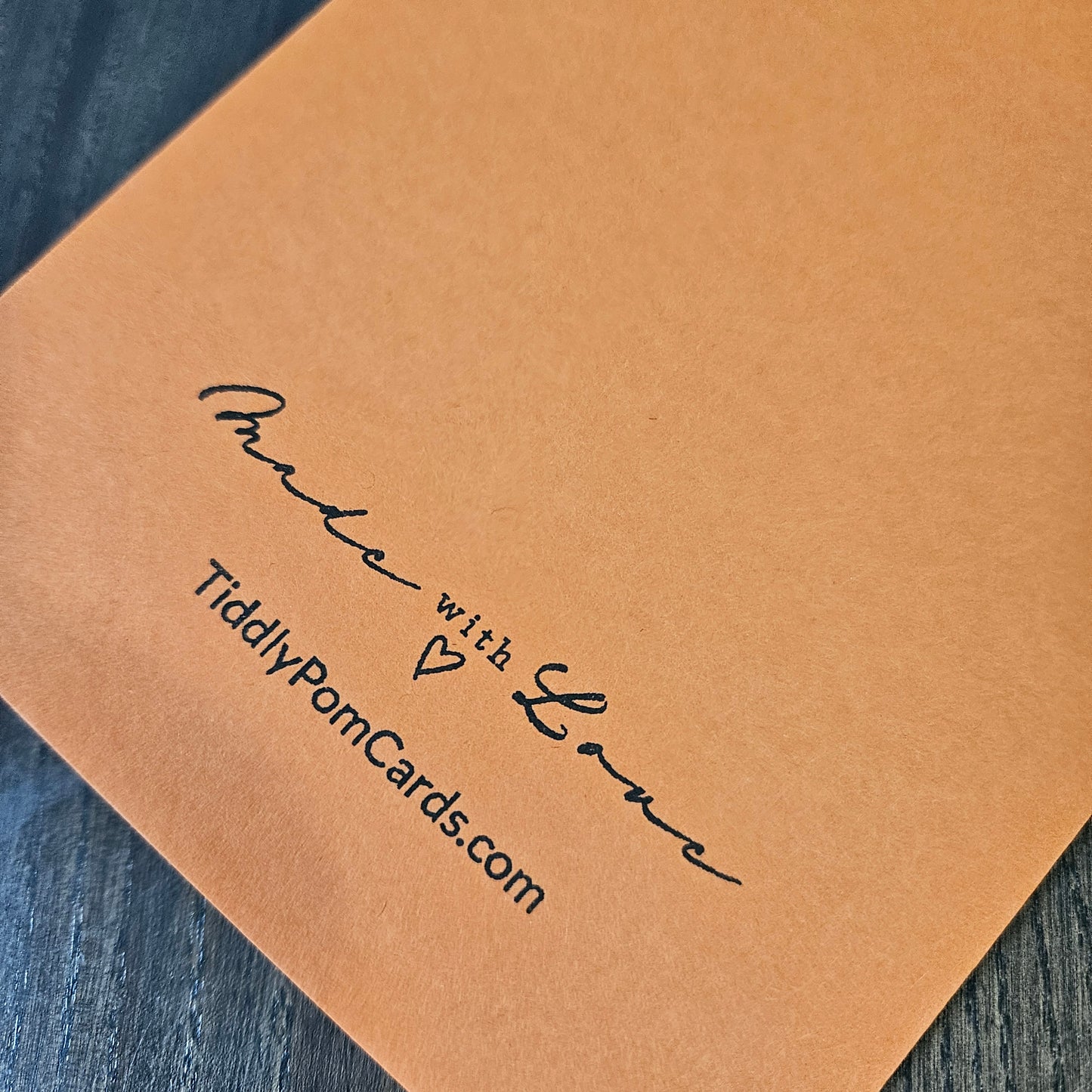 A close-up of a tan kraft paper card or envelope corner printed with the words 'Make with Love' and 'TiddlyPomCards.com' in black script, placed on a dark wood surface.