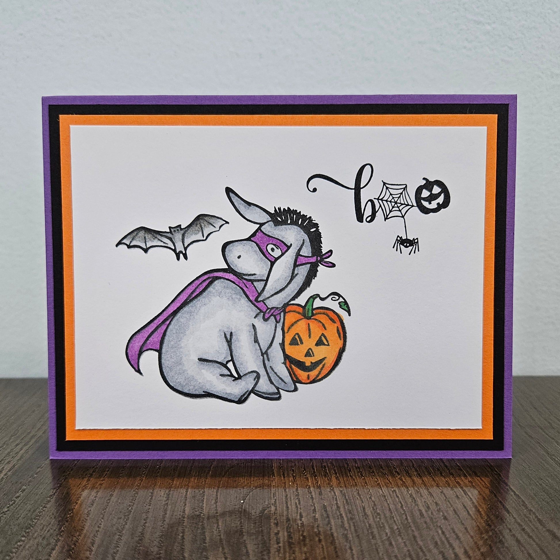 Front of a handmade A2 Halloween card featuring a hand-colored Eeyore in a purple superhero cape and mask next to a pumpkin with a flying bat and Boo sentiment.