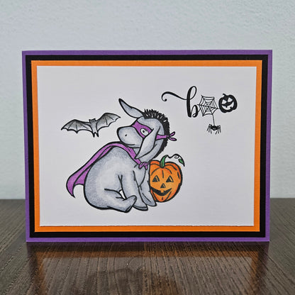 Front of a handmade A2 Halloween card featuring a hand-colored Eeyore in a purple superhero cape and mask next to a pumpkin with a flying bat and Boo sentiment.