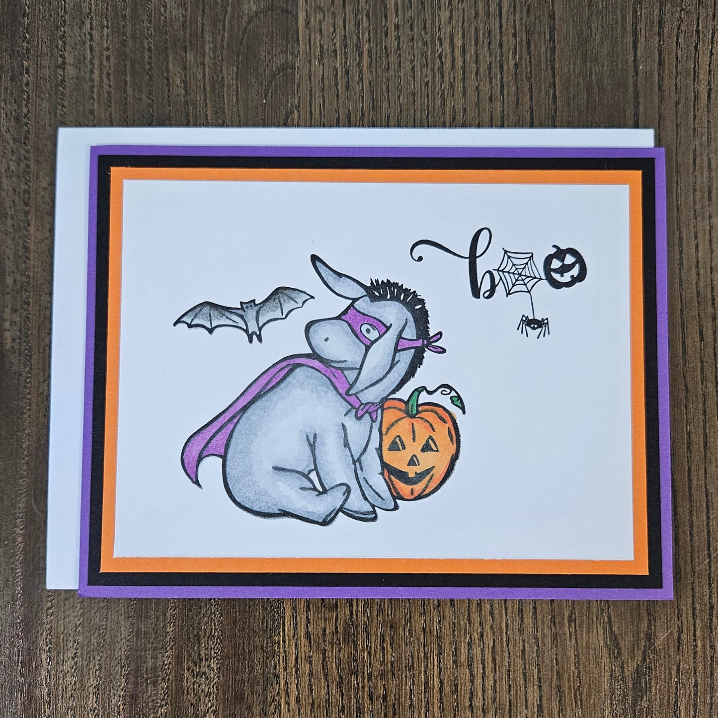 Front of a handmade A2 Halloween card featuring a hand-colored Eeyore in a purple superhero cape and mask next to a pumpkin with a flying bat and Boo sentiment.
