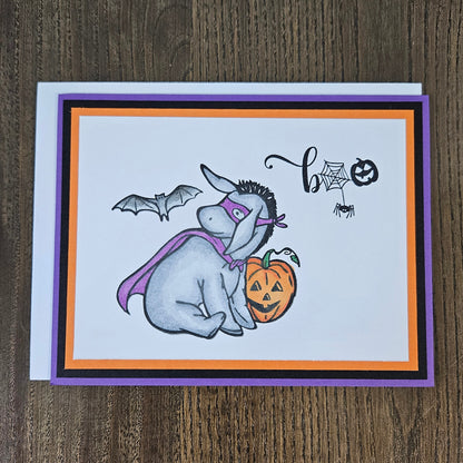 Front of a handmade A2 Halloween card featuring a hand-colored Eeyore in a purple superhero cape and mask next to a pumpkin with a flying bat and Boo sentiment.