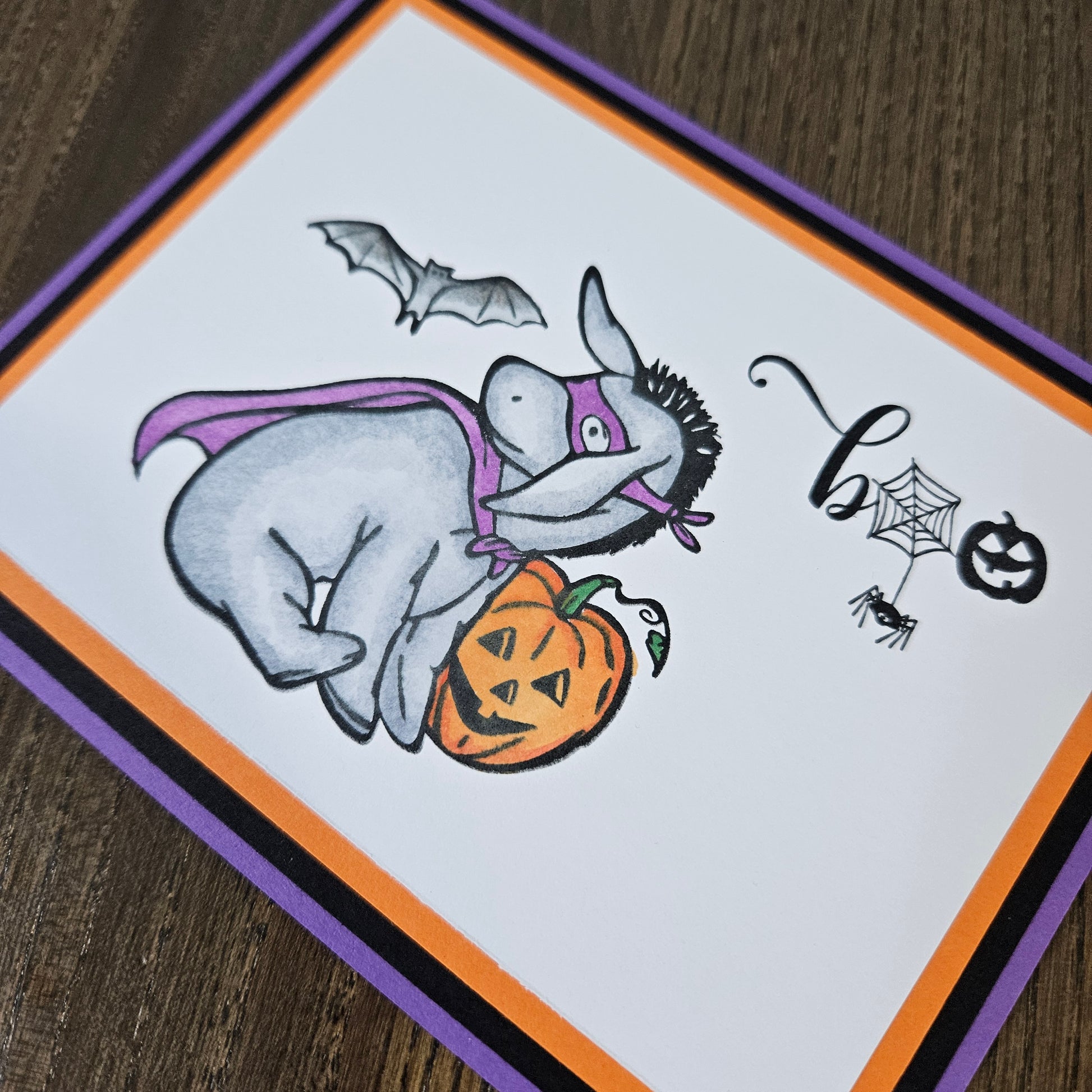 A square Halloween greeting card featuring a cartoon donkey in a purple cape and mask, holding a jack-o'-lantern, with a bat, spiderweb, spider, and jack-o'-lantern on a white card. The card has an orange border and is photographed on a dark wood surface.