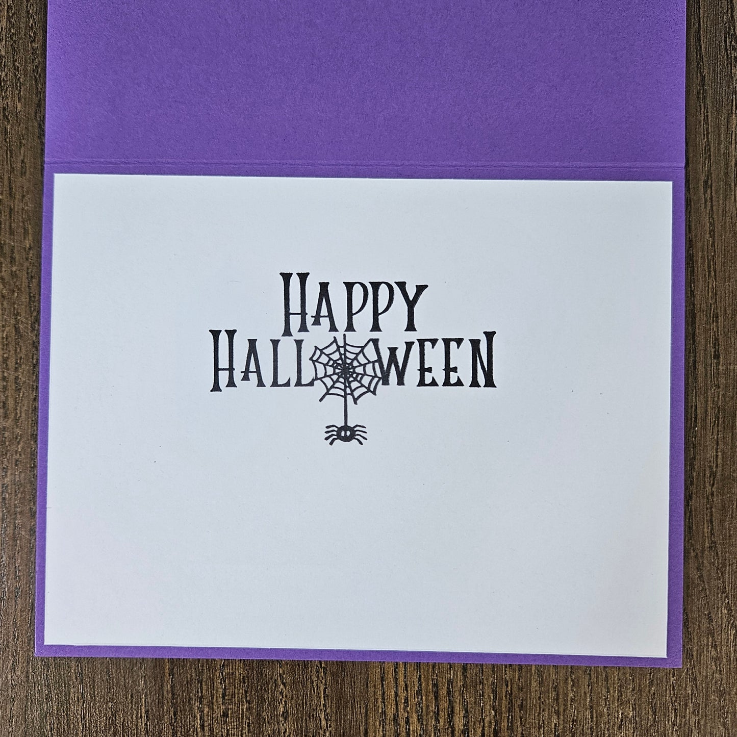 A square purple greeting card with a white front insert printed with 'Happy Halloween' in black decorative lettering and a spiderweb motif, photographed on a wood surface.