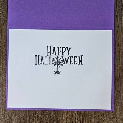 A square purple greeting card with a white front insert printed with 'Happy Halloween' in black decorative lettering and a spiderweb motif, photographed on a wood surface.