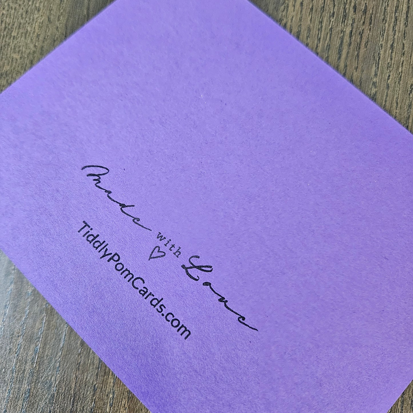 A close-up of a lavender purple textured card with black printed text reading 'Make with Love' and 'TiddlyPomCards.com', placed on a wooden surface.