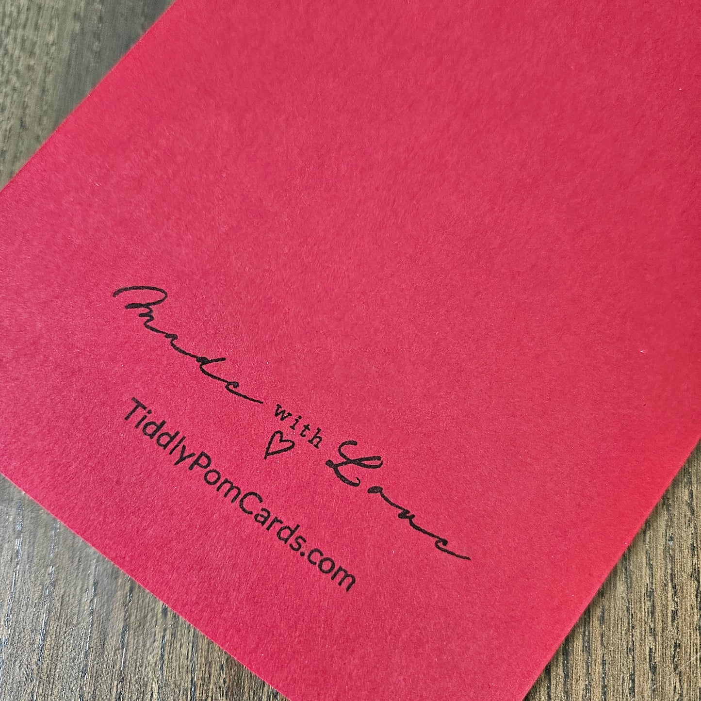 A close-up of a red textured card or paper product placed on a wood surface, printed with the phrase 'Made with Love' and the website 'TidlyPornCards.com' in black script.