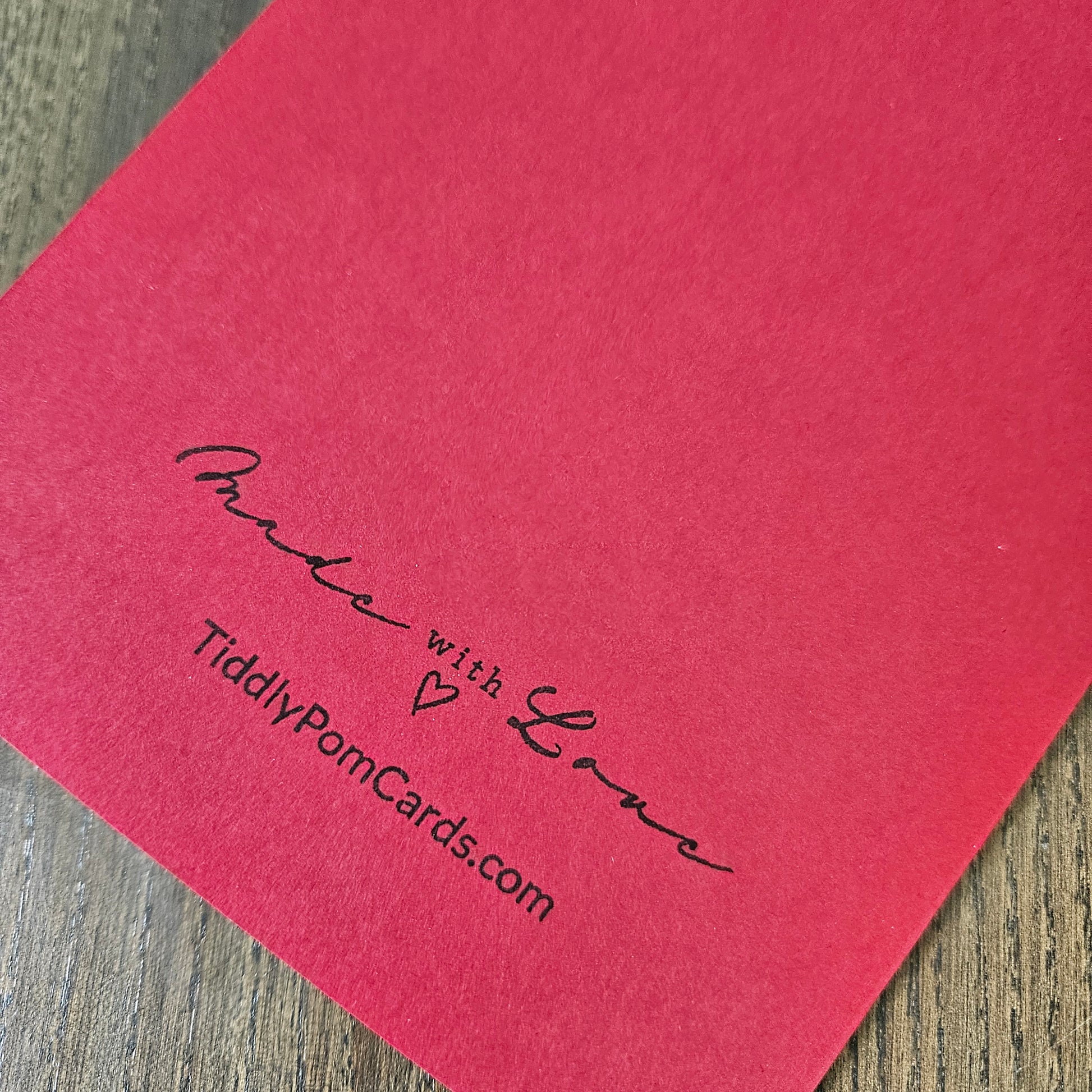 A close-up of a red textured card or paper product placed on a wood surface, printed with the phrase 'Made with Love' and the website 'TidlyPornCards.com' in black script.