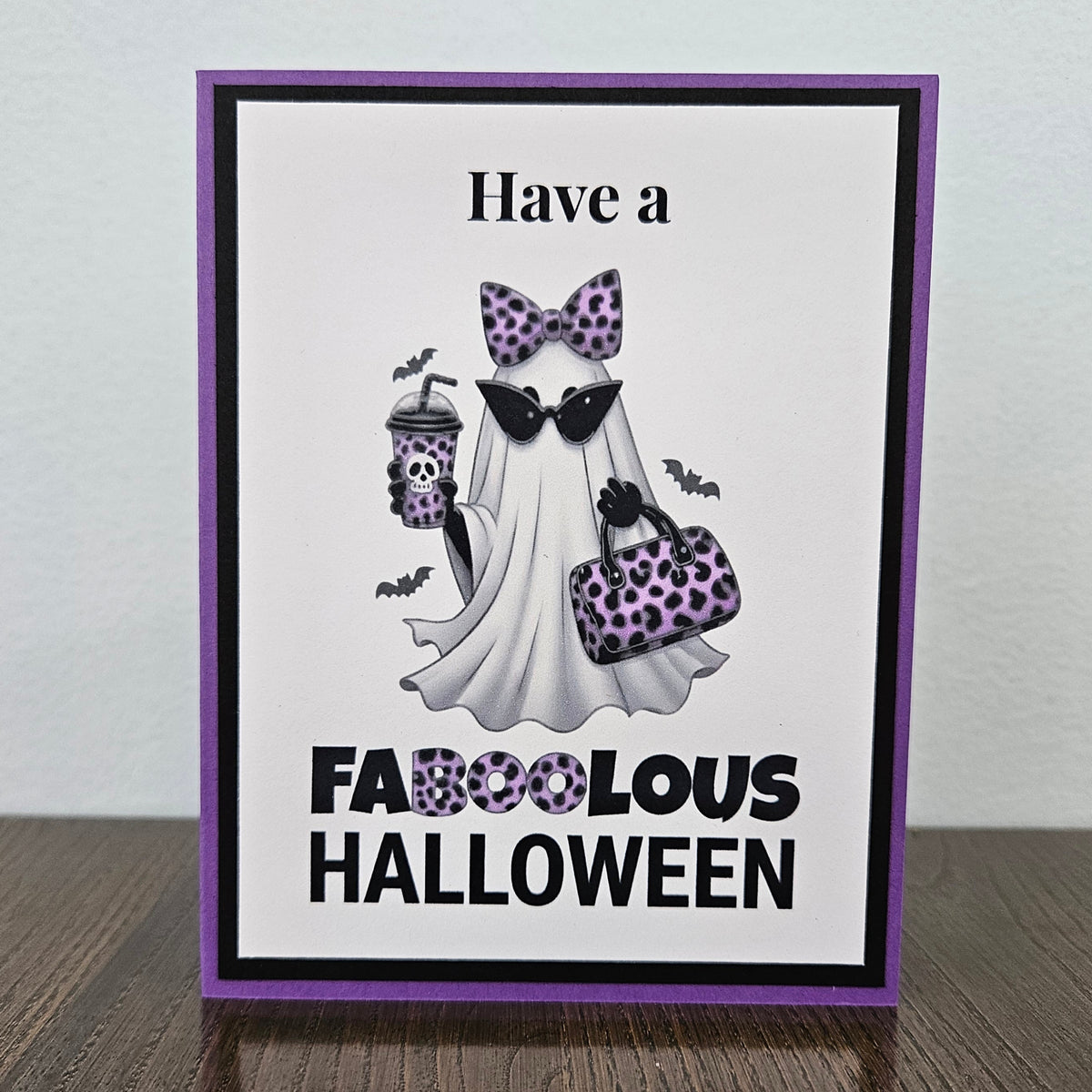 Front of a handmade A2 Halloween card featuring a layered purple and black border around a fashionable ghost wearing a purple leopard bow and sunglasses, holding an iced coffee and purse, with text reading Have a FABOOLOUS HALLOWEEN.