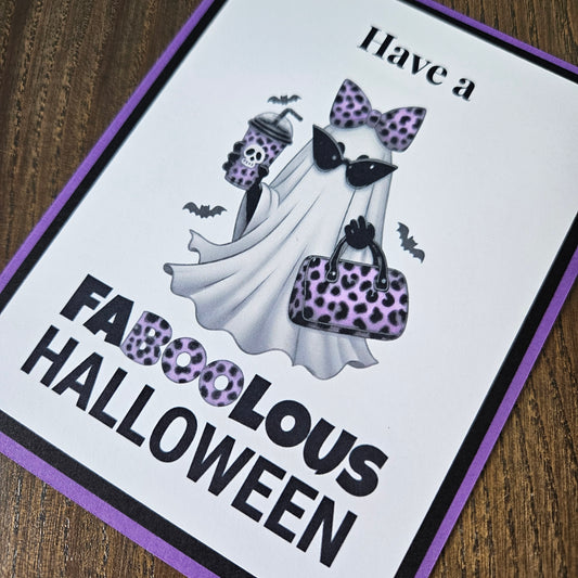 A square printed card or sign with a purple border and the text 'Have a FABULOUS HALLOWEEN' on a wooden surface. The design shows a white ghost wearing a purple-and-black leopard-print bow, sunglasses, and handbag, with small bats and a drink cup.