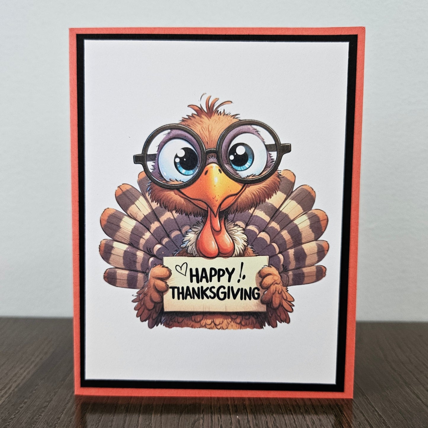 A handmade Thanksgiving greeting card featuring a cartoon turkey wearing round black glasses and holding a sign that reads 'HAPPY THANKSGIVING'. The card has a white center, a black inner border, and a coral-red outer border, standing on a dark wooden surface against a light gray wall.