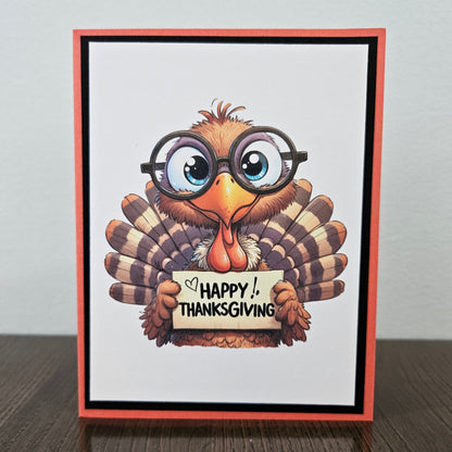 A handmade Thanksgiving greeting card featuring a cartoon turkey wearing round black glasses and holding a sign that reads 'HAPPY THANKSGIVING'. The card has a white center, a black inner border, and a coral-red outer border, standing on a dark wooden surface against a light gray wall.