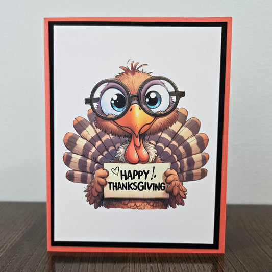A handmade Thanksgiving greeting card featuring a cartoon turkey wearing round black glasses and holding a sign that reads 'HAPPY THANKSGIVING'. The card has a white center, a black inner border, and a coral-red outer border, standing on a dark wooden surface against a light gray wall.