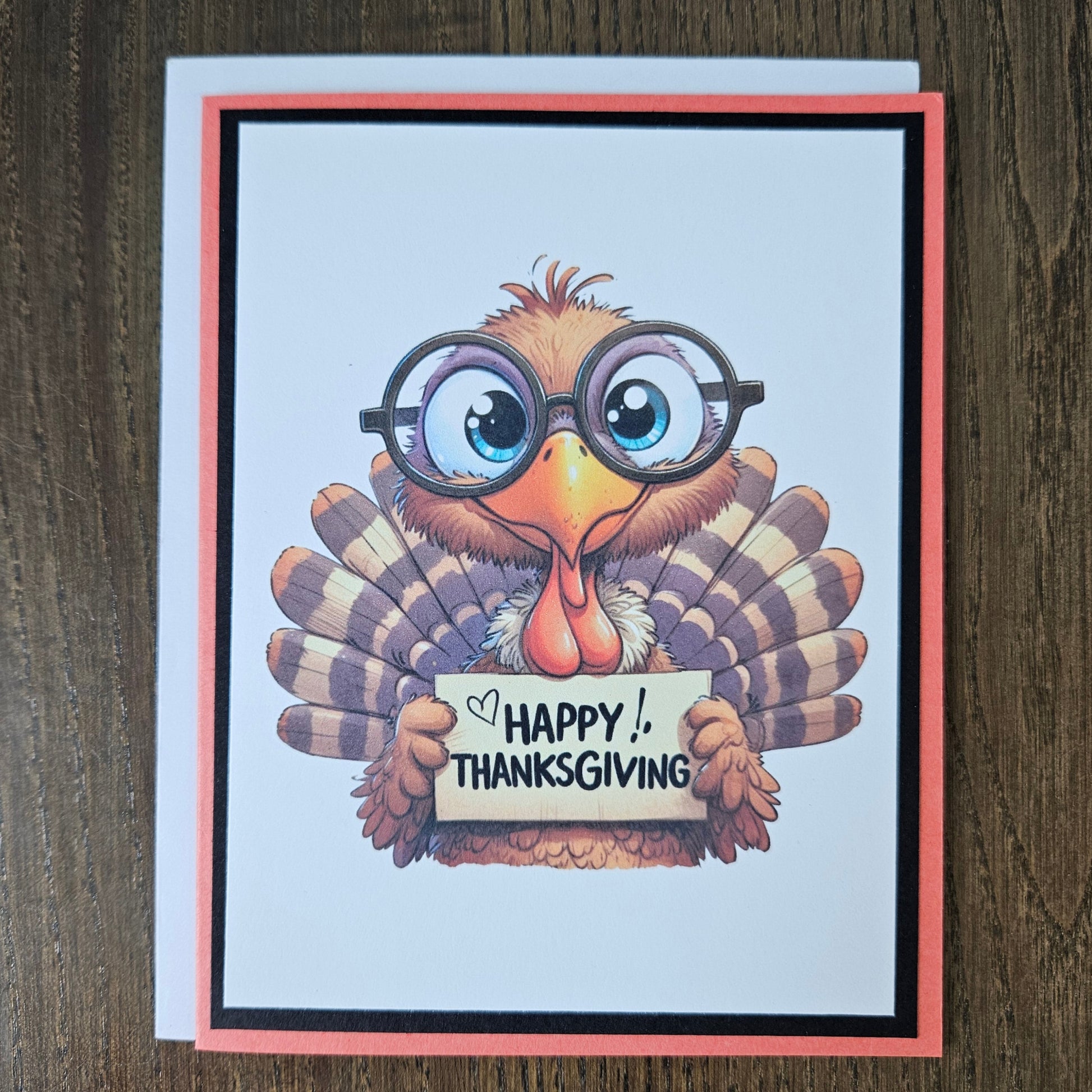 A handmade Thanksgiving greeting card featuring a cartoon turkey wearing round black glasses and holding a sign that reads 'HAPPY THANKSGIVING'. The card has a white background with a black and coral border, placed on a dark wood surface.