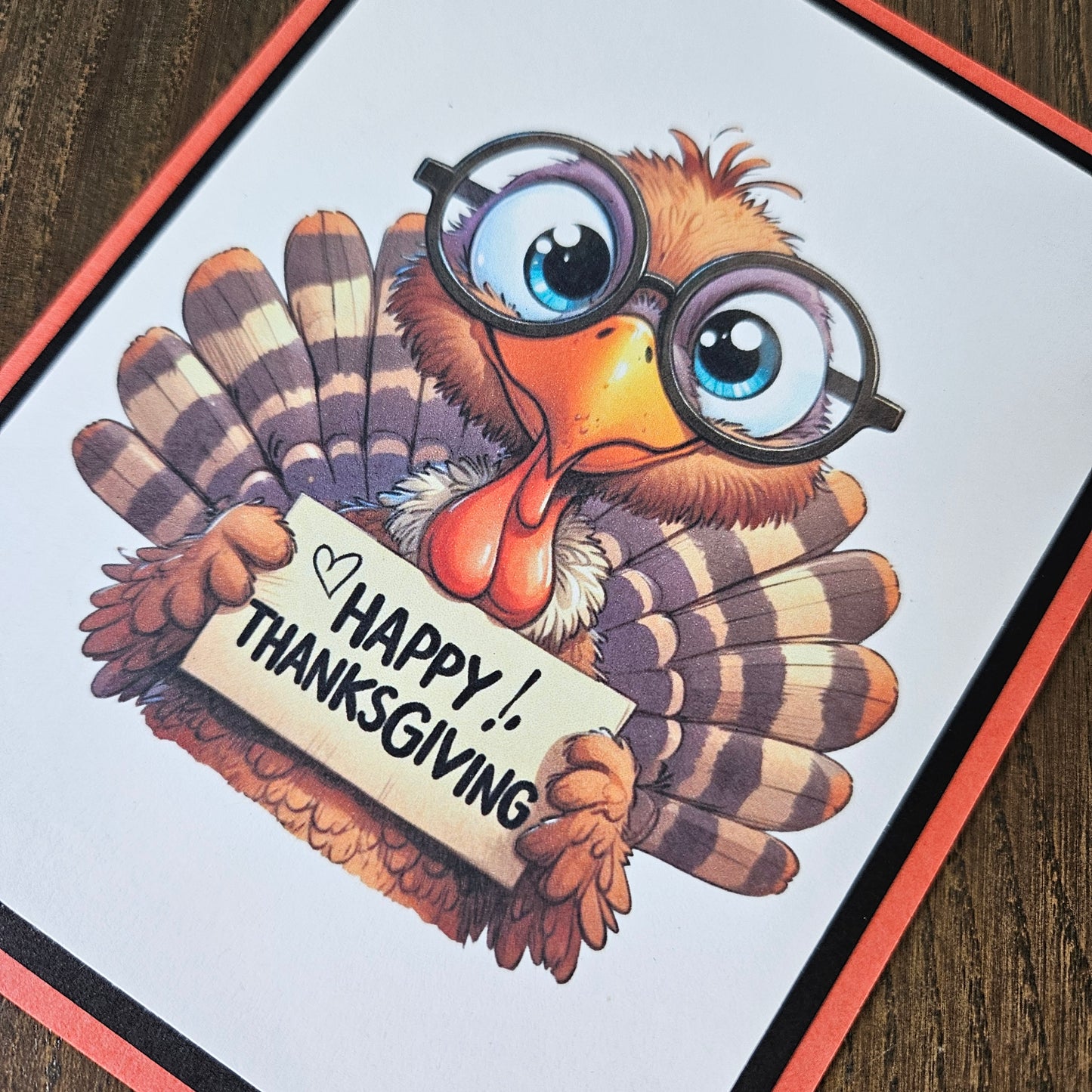 A square greeting card featuring a cartoon turkey with round glasses and a sign that reads 'Happy Thanksgiving,' photographed on a dark wood surface.
