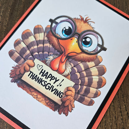 A square greeting card featuring a cartoon turkey with round glasses and a sign that reads 'Happy Thanksgiving,' photographed on a dark wood surface.