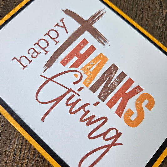 A square white Thanksgiving greeting card or sign with the words “happy Thanks Giving” in brown, orange, and red lettering, bordered in yellow and black, placed on a dark wood surface.