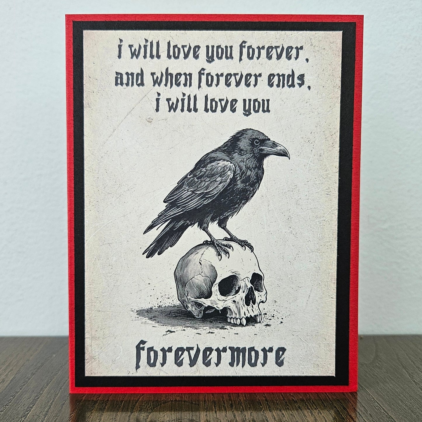A gothic-style art print greeting card featuring a black raven perched on a skull, with the text 'i will love you forever, and when forever ends, i will love you forevermore' in black gothic lettering. The print has a cream textured center, bordered by black and a red outer border, standing on a dark wooden surface against a light gray wall.