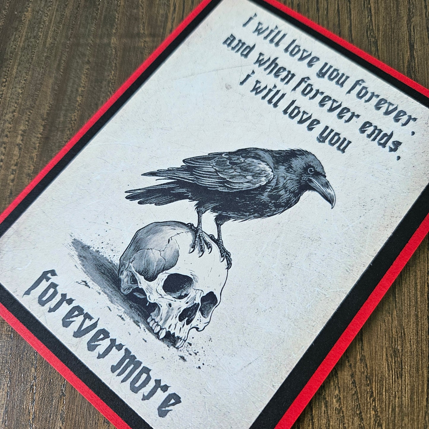 A square art print or card featuring a black raven perched on a skull, with gothic text reading 'I will love you forever and when forever ends, i will love you forevermore,' shown on a wood surface with a red and black border.