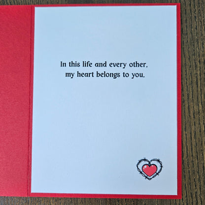 An open red greeting card with a white center panel printed with the phrase 'In this life and every other, my heart belongs to you.' and a small red heart outlined in black at the bottom right, placed on a dark wood surface.