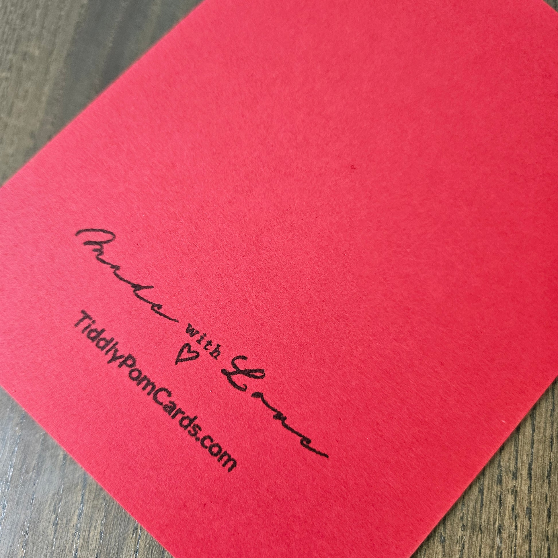 A close-up of a red textured card printed with black cursive text reading 'Purse with Love' and 'TiddlyPornCards.com' on a wooden surface.