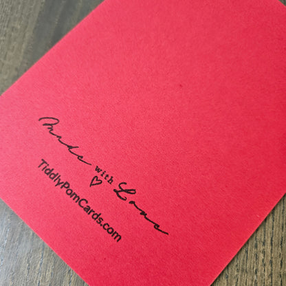 A close-up of a red textured card printed with black cursive text reading 'Purse with Love' and 'TiddlyPornCards.com' on a wooden surface.
