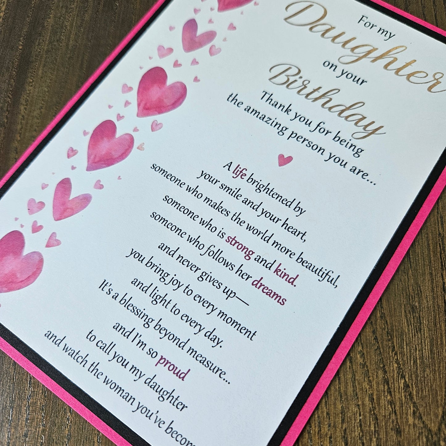A close-up of a printed birthday card or gift message with a white background, pink border, pink watercolor hearts, and cursive and serif text addressed to a daughter. The card is photographed on a dark wood surface.