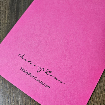 A bright pink textured card or envelope corner placed on a dark wood surface, printed with the words 'Praise with Love' and 'TiddlyPomCards.com' in black script.