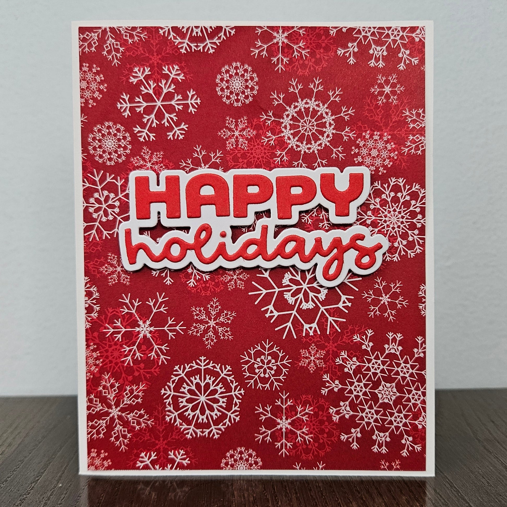 A handmade holiday greeting card with a red background, white snowflake patterns, and a layered 'HAPPY holidays' text design, standing on a dark wooden surface against a light gray wall.