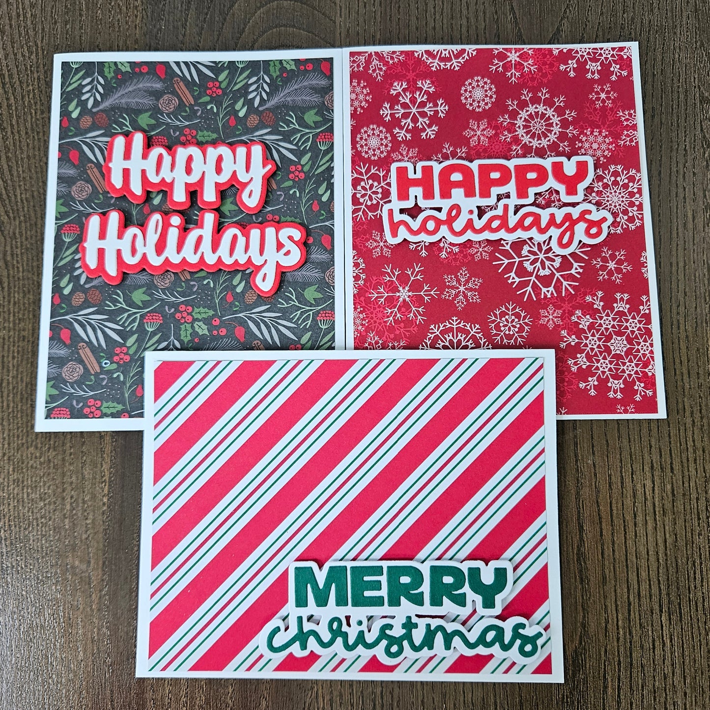 Three handmade Christmas greeting cards arranged on a dark wood background. One card has a dark green holiday pattern with red berries and white 'Happy Holidays' text.