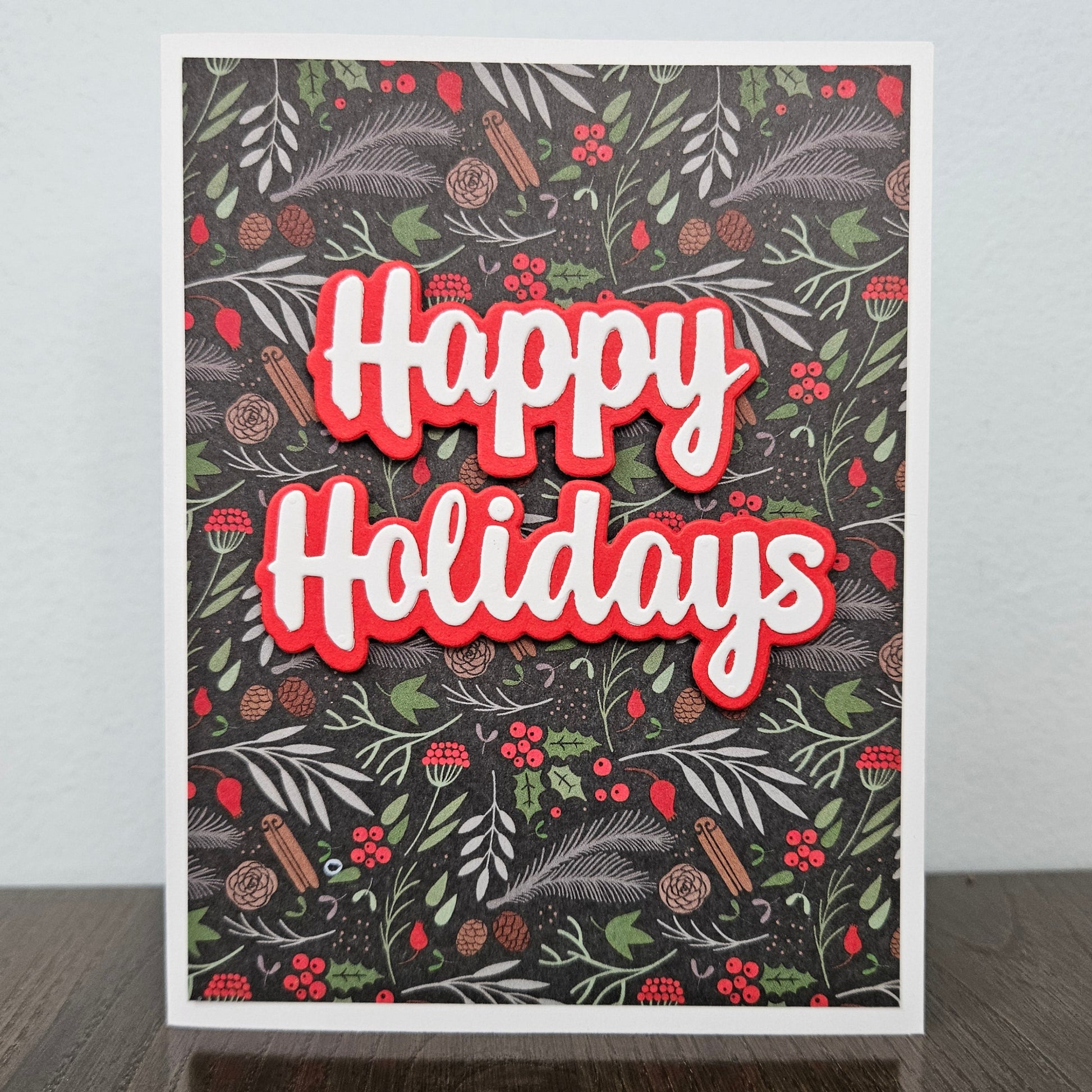 A square holiday greeting card standing upright on a dark wooden surface against a light gray wall. The card features a dark patterned background with green foliage, red berries, pinecones, and cinnamon sticks, with large layered 'Happy Holidays' text in white with a red outline.