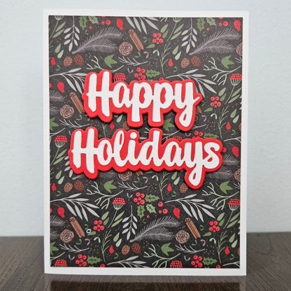 A square holiday greeting card standing upright on a dark wooden surface against a light gray wall. The card features a dark patterned background with green foliage, red berries, pinecones, and cinnamon sticks, with large layered 'Happy Holidays' text in white with a red outline.