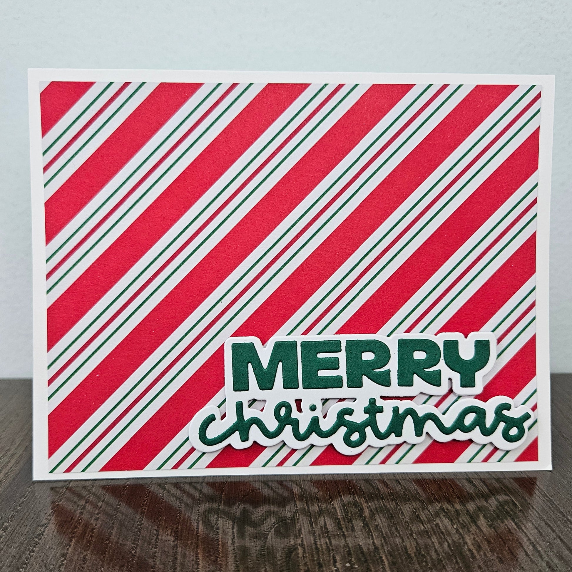 A square Christmas greeting card with diagonal red, white, and green stripes and the words “MERRY christmas” in green, standing on a dark wooden surface against a light gray wall.