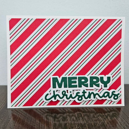 A square Christmas greeting card with diagonal red, white, and green stripes and the words “MERRY christmas” in green, standing on a dark wooden surface against a light gray wall.
