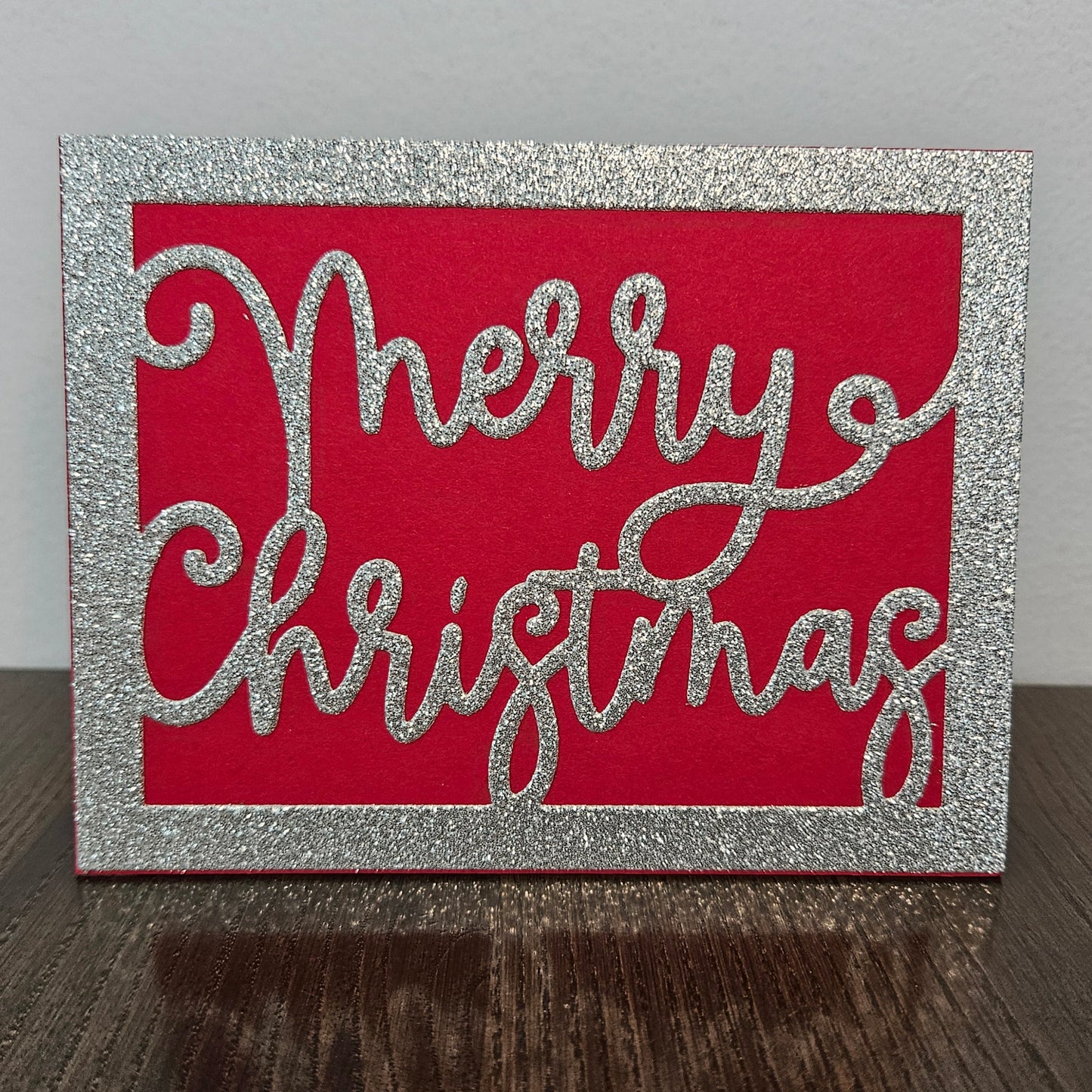 A square Christmas card with a glittery silver border and the words 'Merry Christmas' in glittery silver script over a bright red background, standing on a dark wooden surface against a light gray wall.