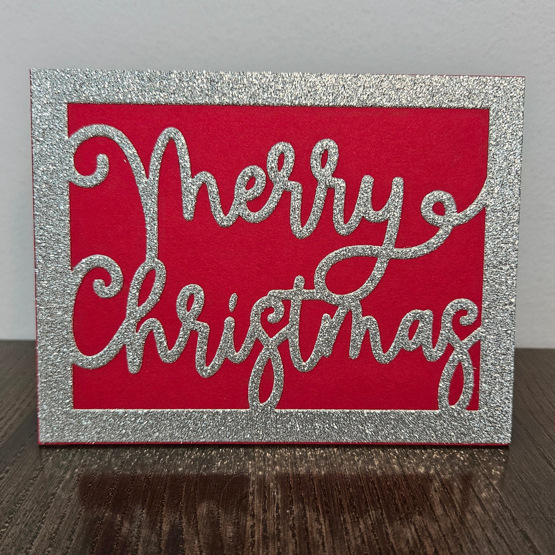 A square Christmas card with a glittery silver border and the words 'Merry Christmas' in glittery silver script over a bright red background, standing on a dark wooden surface against a light gray wall.