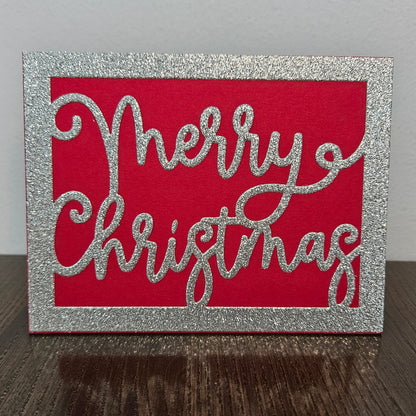 A square Christmas card with a glittery silver border and the words 'Merry Christmas' in glittery silver script over a bright red background, standing on a dark wooden surface against a light gray wall.