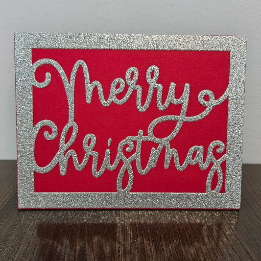 A square Christmas card with a glittery silver border and the words 'Merry Christmas' in glittery silver script over a bright red background, standing on a dark wooden surface against a light gray wall.