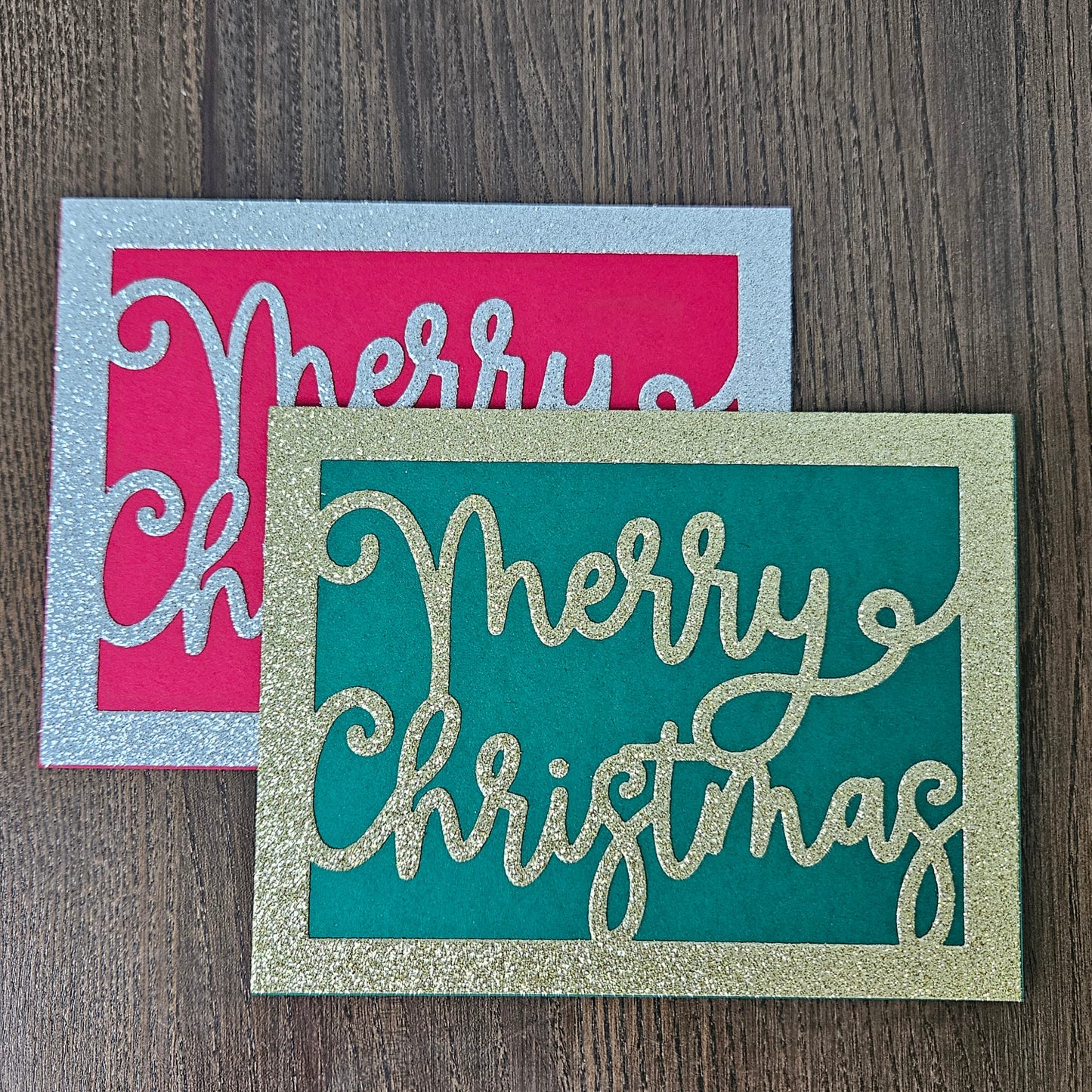 Two decorative holiday cards on a dark wood surface. The front card is teal with glittery gold 'Merry Christmas' lettering and a glittery gold border.