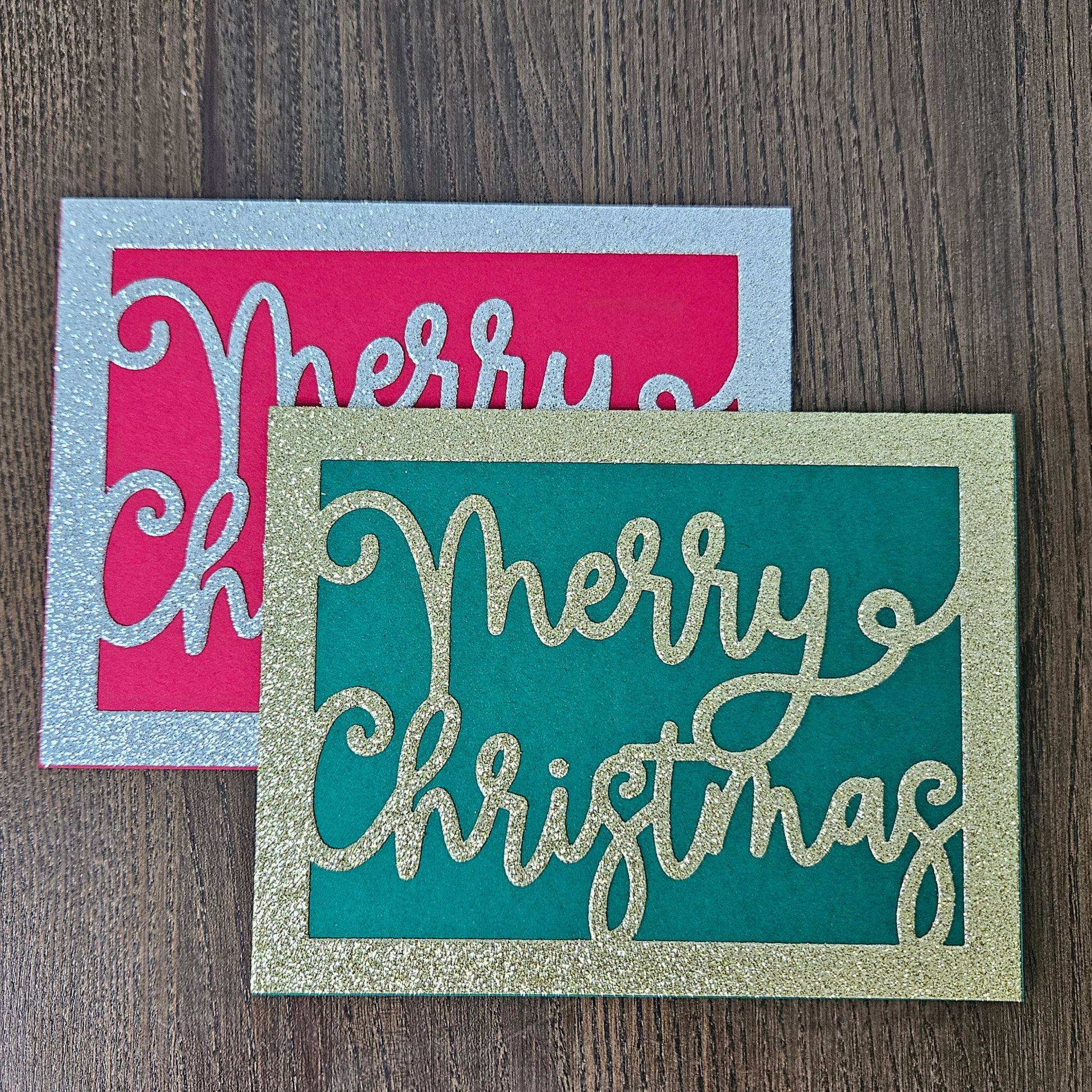 Two decorative holiday cards on a dark wood surface. The front card is teal with glittery gold 'Merry Christmas' lettering and a glittery gold border.