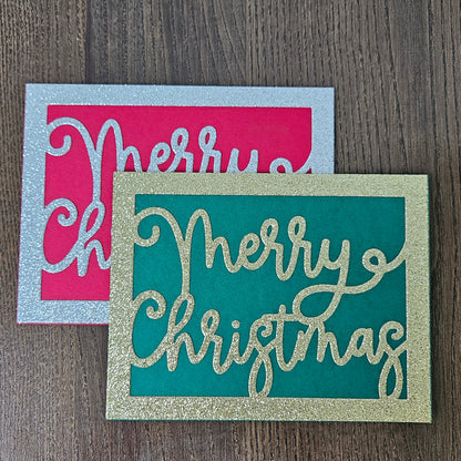 Two decorative holiday cards on a dark wood surface. The front card is teal with glittery gold 'Merry Christmas' lettering and a glittery gold border.