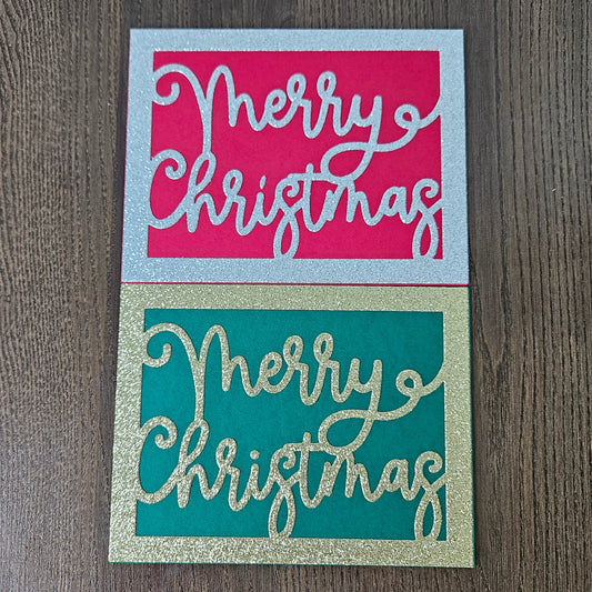 Two square Christmas greeting cards on a dark wood background. The top card has a silver glitter border with a bright pink center and white cursive text reading 'Merry Christmas.
