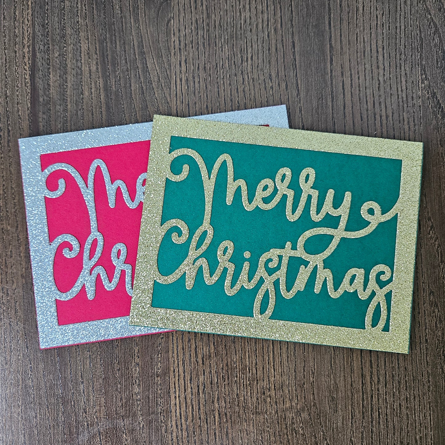 Two square Christmas greeting cards with glitter borders, displayed on a dark wood background. One card has a teal background with gold glitter script reading 'Merry Christmas', and the other has a pink background with a silver glitter border and partial script text.