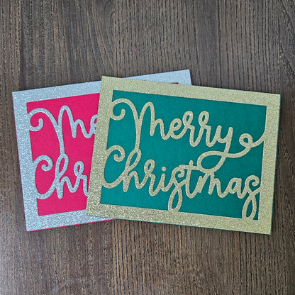 Two square Christmas greeting cards with glitter borders, displayed on a dark wood background. One card has a teal background with gold glitter script reading 'Merry Christmas', and the other has a pink background with a silver glitter border and partial script text.