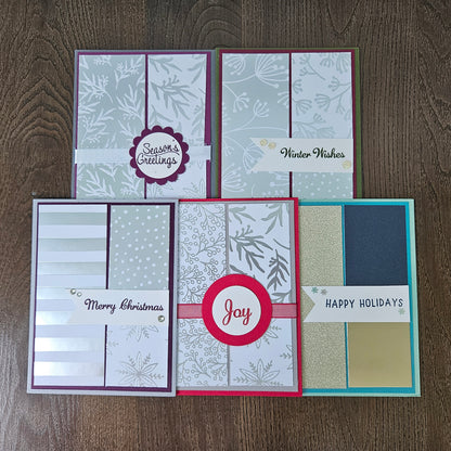Five handmade Christmas greeting cards arranged on a dark wood background. The cards feature various holiday patterns, colors, and sentiments including 'Season's Greetings', 'Winter Wishes', 'Merry Christmas', 'Joy', and 'Happy Holidays'.
