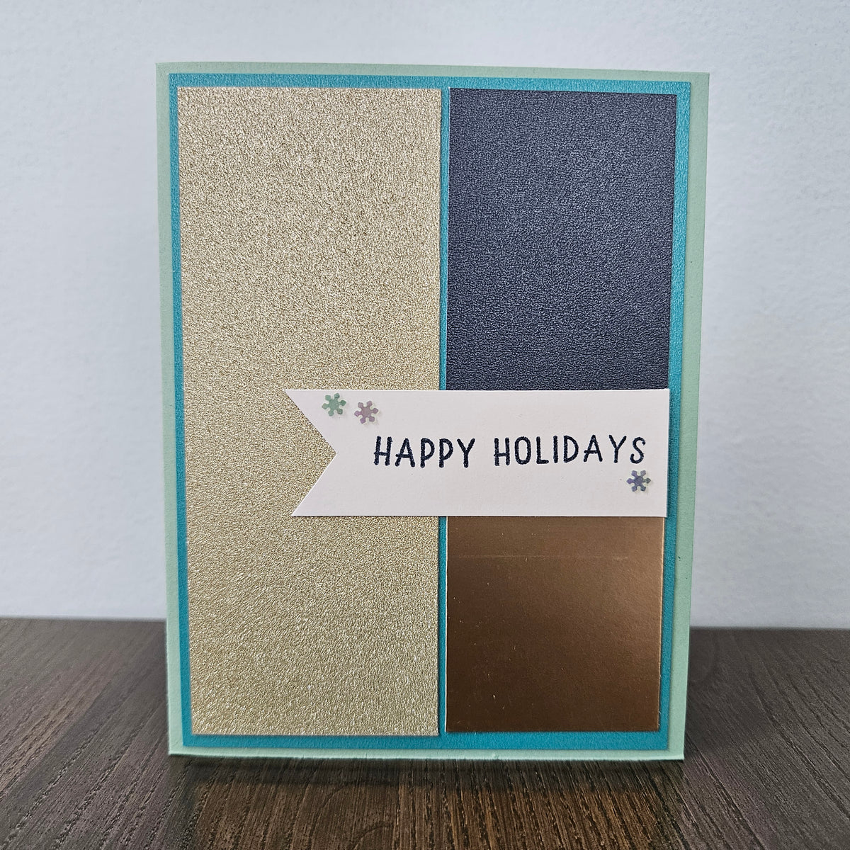 A handmade holiday greeting card with a teal border, featuring four vertical panels in glittery gold, dark blue, metallic bronze, and white, with a banner reading 'HAPPY HOLIDAYS' on a wooden surface.