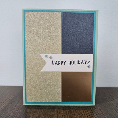 A handmade holiday greeting card with a teal border, featuring four vertical panels in glittery gold, dark blue, metallic bronze, and white, with a banner reading 'HAPPY HOLIDAYS' on a wooden surface.