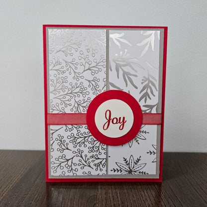 A handmade greeting card with a red border, four patterned white and silver panels, a red ribbon across the center, and a circular red and white badge reading 'Joy' in red script, standing on a dark wooden surface against a light gray wall.