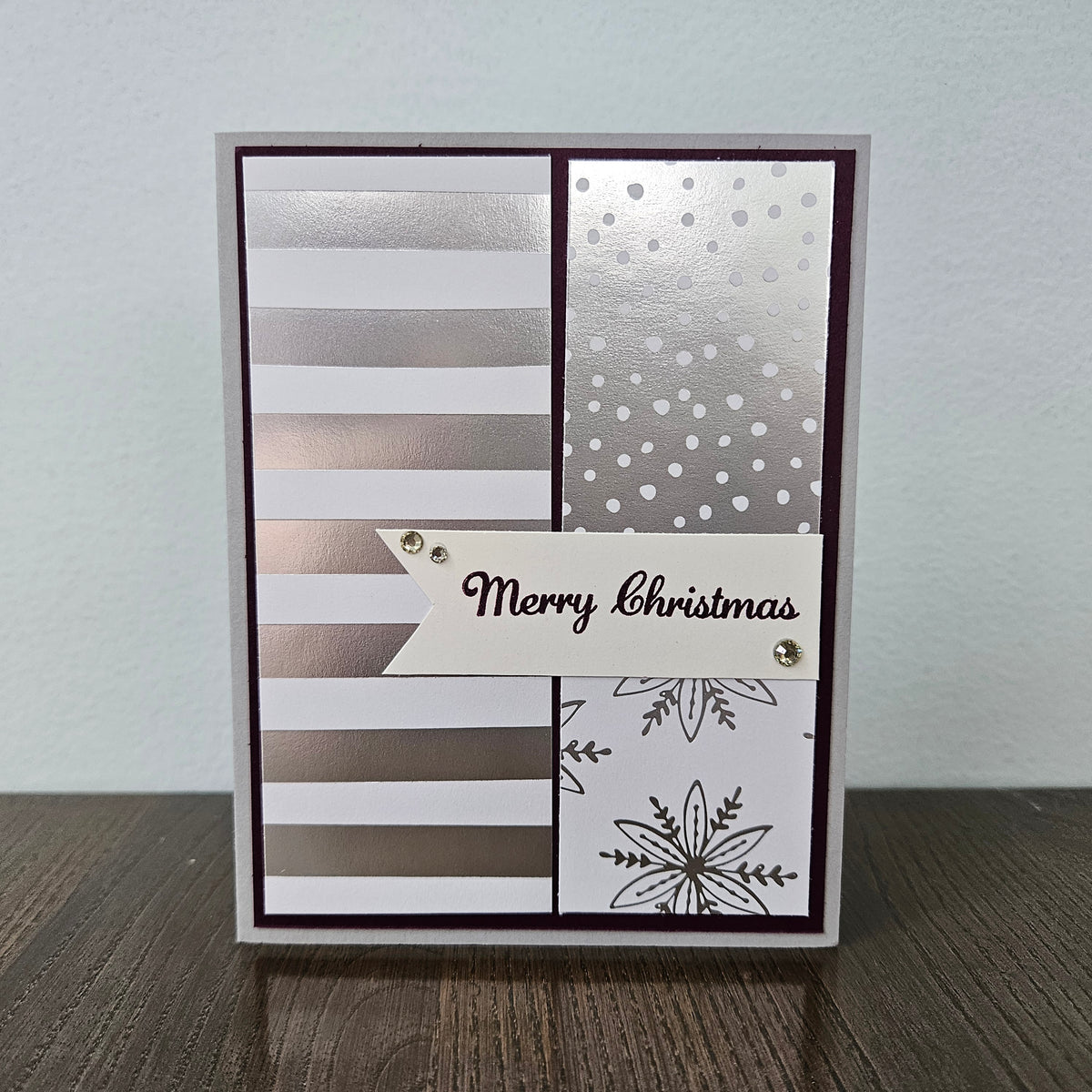 A handmade Christmas greeting card with a silver and white striped and polka dot background, featuring a white banner that reads 'Merry Christmas' and a black snowflake design at the bottom, displayed on a wooden surface against a light gray wall.
