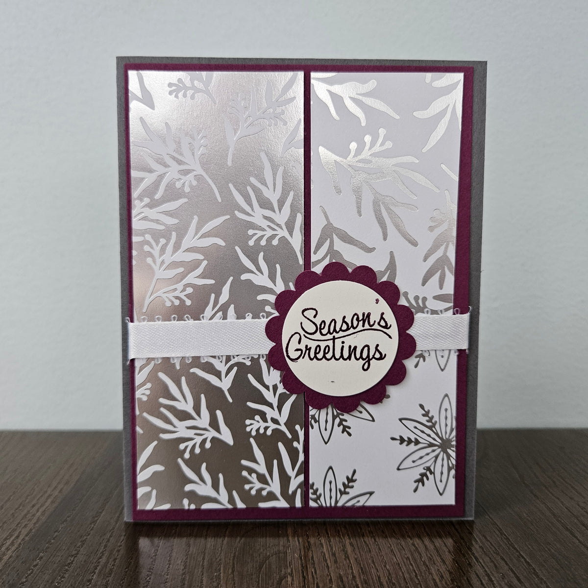 A handmade greeting card with a silver and white botanical pattern, featuring a central scalloped burgundy circle with the words 'Season's Greetings' and a white ribbon across the middle, displayed on a wooden surface against a light gray background.